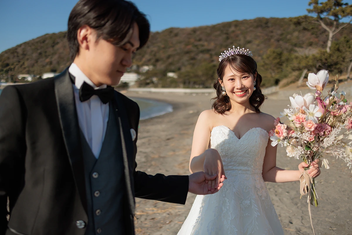 タキシードを着た新郎とウェディングドレスを着た新婦がブーケを持ち海沿いを歩くウェディングフォト