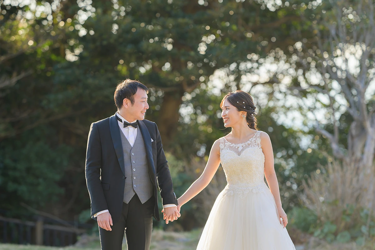 タキシードを着た新郎とラウンドネックのウェディングドレスを着た新婦が手をつないでほほ笑む前撮りフォト