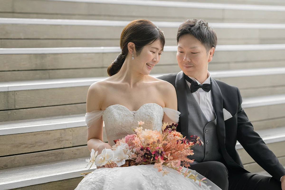 大桟橋で洋装の新郎新婦がブーケを持ち座ってほほ笑むウェディングフォト