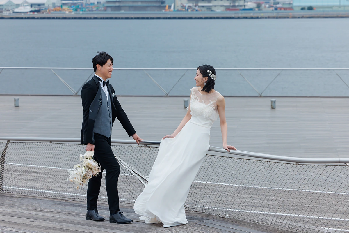 大桟橋でスレンダーラインのドレスを着る新婦とタキシードを着た新郎が見つめ合うウェディングフォト