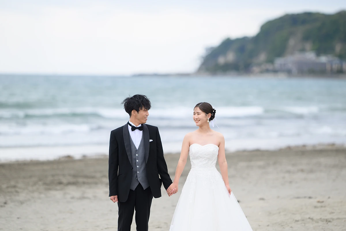 海辺でウェディングドレスを着た新婦とタキシードを着用した新郎が手を繋いでほほ笑む洋装ウェディングフォト