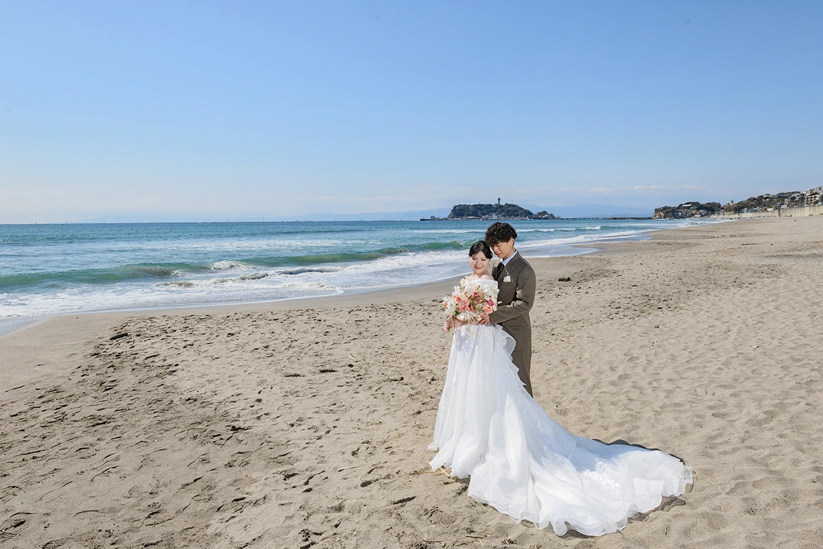 七里ガ浜でウェディングドレスとタキシードを着用した新郎新婦が江ノ島を背景に写る洋装前撮り