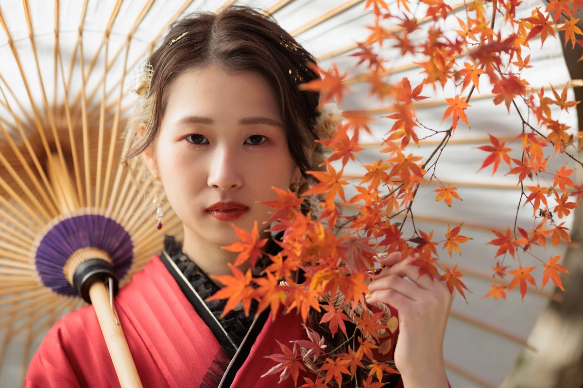 紅葉と和傘を背景に、赤系の振袖姿でカメラを見つめる女性の上半身を捉えた鎌倉の成人式前撮り。