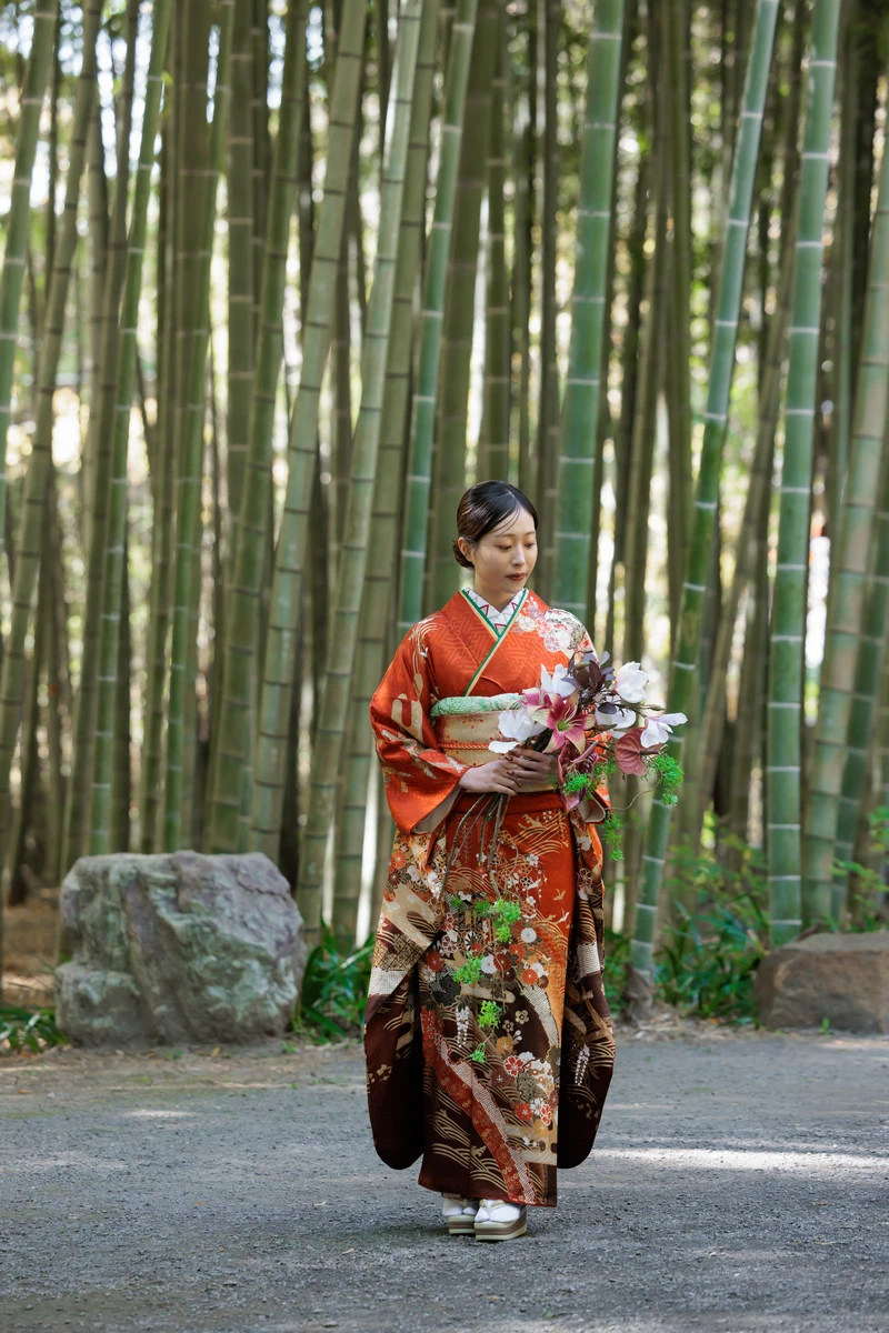 鎌倉建長寺の竹林で、橙色と茶系の振袖姿の女性がブーケを両手に持って立つ成人式前撮りの全身カット。