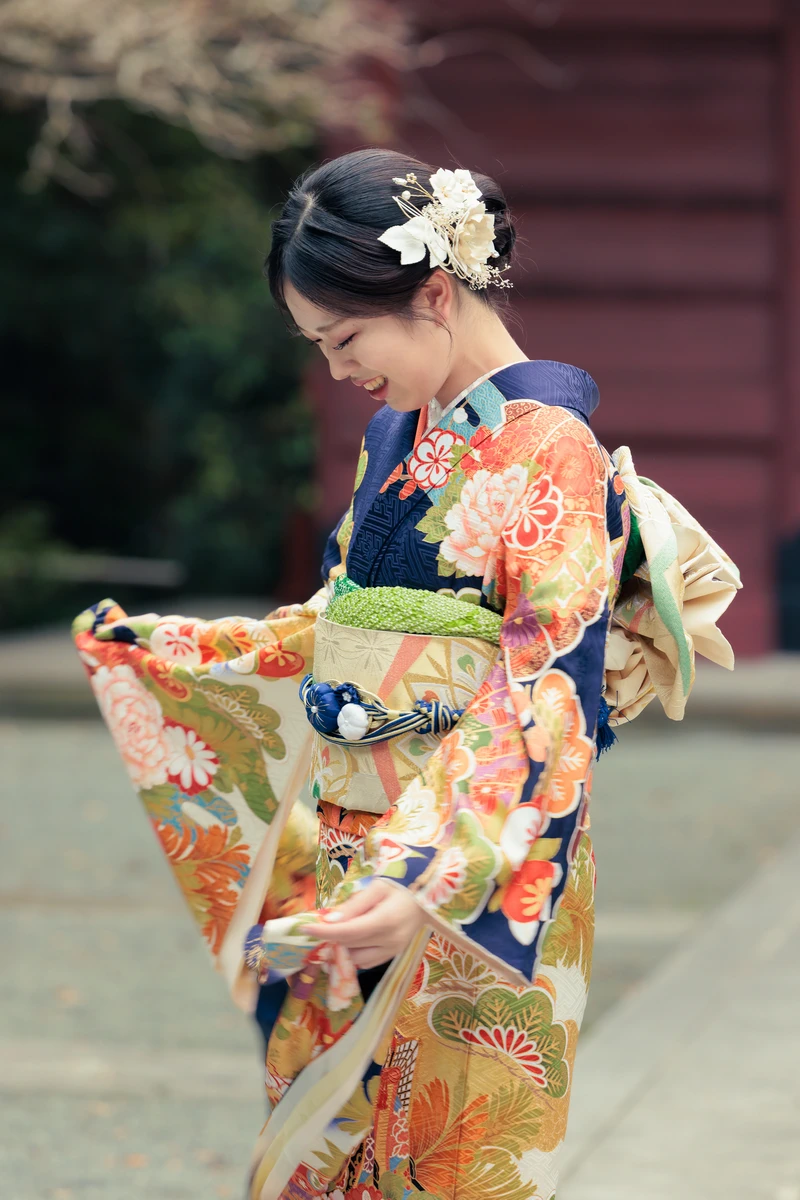 紺地に赤や金の花文様が入った振袖姿の女性が、白い髪飾りと帯結びを見せながら微笑む成人式前撮り。