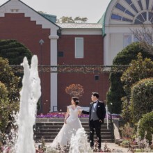 噴水越しに見つめ合う洋装の新郎新婦と赤レンガの洋館