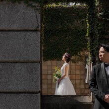 石壁越しに振り返るタキシード姿の新郎と、緑に囲まれた通路に佇むウェディングドレス姿の新婦