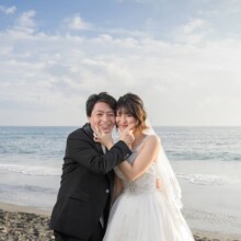 七里ヶ浜の海辺で頬を寄せ合い笑顔を見せる新郎新婦。青い海を背景にした洋装フォトウェディングの記念カット。