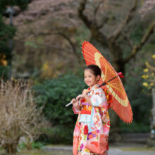 赤い和傘を肩に担いで歩く晴れ着姿の女の子。妙本寺の自然豊かな境内で撮影した七五三の記念ポートレート。