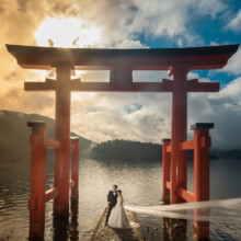 夕暮れの中湖の上に立つ鳥居と、鳥居の前に並ぶ洋装の新郎新婦 女性のベールが風になびいている