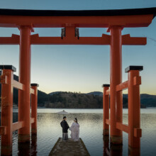 朝焼けの中湖の上に立つ鳥居と、鳥居の前に並ぶ和装の新郎新婦