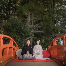森の中にかかる朱色の橋と、橋の上で正座して並ぶ和装の新郎新婦 和傘が添えられている