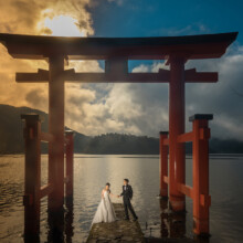 夕暮れの中湖の上に立つ鳥居と、鳥居の前に並ぶ洋装の新郎新婦