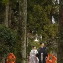 森の中にかかる朱色の橋と、手を取り合い渡る和装の新郎新婦