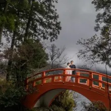 湖畔の森に佇む湖をまたぐ朱色の橋と、その上で並ぶ和装の新郎新婦