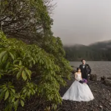 木の生い茂る湖畔で並ぶ洋装の新郎新婦 女性はブーケを持っている
