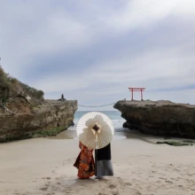 伊豆 和装 K様・Y様 伊豆の砂浜で白い番傘を差す色打掛の花嫁と黒紋付袴の新郎が、海辺の岩上に立つ赤い鳥居を望む和装前撮り