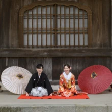 鎌倉 和装 K様・S様 2026.02.24 鎌倉建長寺の木造壁を背景に、赤い毛氈で正座する色打掛の花嫁と黒紋付袴の新郎を、紅白の和傘が囲む和装前撮り