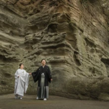 鎌倉 和装 K様・K様 2020.03.31 英勝寺の切り立った岩壁の前で、白無垢の新婦と黒紋付袴の新郎が手をつないで立つ広角構図