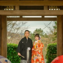 鎌倉 和装 N様・M様 2021.03.22 海蔵寺の山門前で、オレンジの色打掛姿の新婦と黒紋付袴姿の新郎が並び、足元に紫と赤の和傘を置く鎌倉前撮り