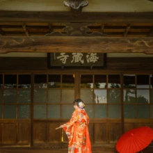 鎌倉 和装 N様・M様 2021.03.22 海蔵寺の本堂前で、オレンジの色打掛姿の新婦が末広を手に横向きに立ち、両側に紫と赤の和傘を添える
