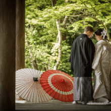 鎌倉 和装 S様・S様 2021.04.02 新緑に囲まれた縁側で、白無垢の新婦と黒紋付袴の新郎が額を寄せ合い、足元に赤と白の和傘を置く
