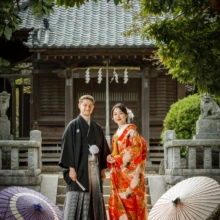 鎌倉 和装 S様・S様 2021.04.02 新緑に囲まれた神社の拝殿前で、赤い色打掛の新婦と黒紋付袴の新郎が三本の和傘と並んで立つ