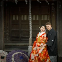 鎌倉 和装 S様・S様 2021.04.02 神社の本殿前で、赤い色打掛の新婦と黒紋付袴の新郎が寄り添い、足元に白と紫の和傘を並べた和装前撮り