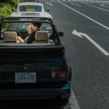 東京 洋装 N様・N様 2021.04.22 都内の道路を走る黒いオープンカーの後部座席から、前席の新郎に笑顔で寄り添うウェディングドレス姿の新婦