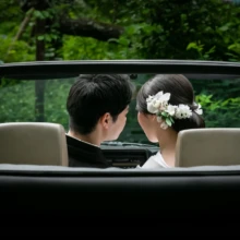 鎌倉 和装 I様・S様 2021.05.28 新緑の中を走る黒いオープンカーの後部座席で、寄り添って座る白無垢の新婦と黒紋付袴の新郎の後ろ姿