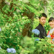 鎌倉 和装 M様・K様 2021.05.30 青い紫陽花と新緑の間から、赤い色打掛の新婦と黒紋付袴の新郎が振り返って微笑む鎌倉フォトウェディング