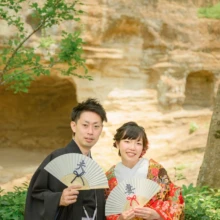鎌倉 和装 M様・K様 2021.05.30 瑞泉寺の岩庭を背景に、扇子を持った赤い色打掛の新婦と黒紋付袴の新郎が並んで微笑む鎌倉和装前撮り
