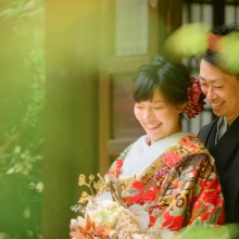 鎌倉 和装 M様・K様 2021.05.30 新緑越しに、花のブーケを持つ赤い色打掛の新婦へ黒紋付袴の新郎が後ろから寄り添う鎌倉前撮り