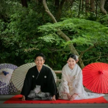鎌倉 和装 O様・M様 2021.05.31 妙本寺の本堂前に敷いた赤い毛氈に正座し、紫と赤の和傘を背に微笑む白無垢の新婦と黒紋付袴の新郎