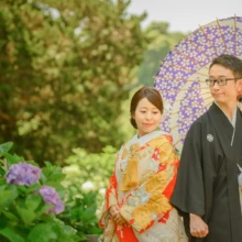 鎌倉 和装 M様・Y様 2021.06.01 建長寺の紫陽花のそばで、紫の和傘を差す黒紋付袴の新郎と朱赤の色打掛の新婦が並ぶ和装前撮り