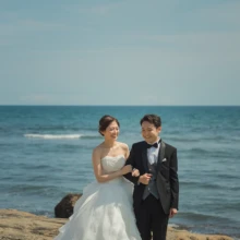 鎌倉 和洋装 H様・S様 2021.06.15 青い海を背景に、ウェディングドレスの新婦がタキシード姿の新郎と腕を組んで岩場を歩く鎌倉フォトウェディング