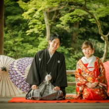 鎌倉 和装 T様・T様 2021.06.21 妙本寺の本堂前に正座し、白・紫・赤の和傘を背に微笑む朱赤の色打掛の新婦と黒紋付袴の新郎の鎌倉和装前撮り