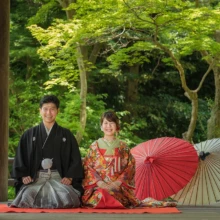 鎌倉 和装 M様・N様 2021.06.28 妙本寺の回廊で、黒紋付袴の新郎と赤い色打掛姿の新婦が正座し、二本の和傘を横に並べた鎌倉和装前撮り