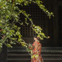 鎌倉 和装 M様・N様 2021.06.28 妙本寺の本堂前で、赤や金の色打掛姿の新婦が枝葉越しに横を向いて立つ鎌倉前撮りの花嫁ソロ