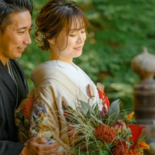 鎌倉 和装 S様・N様 2021.07.12 妙本寺の夏の緑を背景に、黒紋付袴の新郎が赤い花のブーケを持つ淡色の色打掛姿の新婦に寄り添う鎌倉和装前撮り