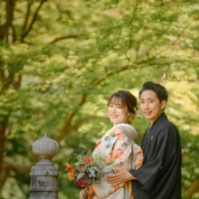 鎌倉 和装 S様・N様 2021.07.12 妙本寺の緑を背景に、赤い花のブーケを抱えた色打掛の新婦へ黒紋付袴の新郎が後ろから寄り添う鎌倉フォトウェディング