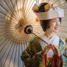 鎌倉 和装 T様・M様 2021.12.20 妙本寺の紅葉を背景に、白い和傘を肩に差して笑顔を見せる角隠しと緑の色打掛姿の新婦