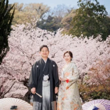 鎌倉 和装 M様・M様 2022.04.01 満開の桜を背景に正面を向いて並ぶ、淡い色打掛の新婦と黒紋付袴の新郎を全身で収めた鎌倉前撮り