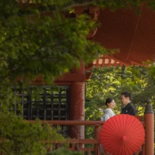 日光 和装 M様・M様 2022.08.29 夏の緑に囲まれた朱塗りの寺院で、赤い和傘を差して向かい合う白無垢姿の新婦と黒紋付姿の新郎