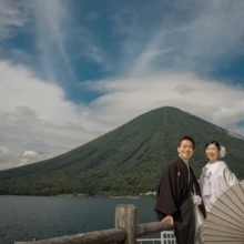 日光 和装 M様・M様 2022.08.29 中禅寺湖と男体山を背景に、黒紋付姿の新郎と白無垢姿の新婦が白い和傘を添えて並ぶ日光和装前撮り