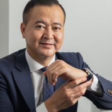 プロフィール写真 I様 2022.09.22 紺色のスーツ姿の男性が白い台に肘を置き、腕時計へ手を添えながらカメラを見る上半身ポートレート