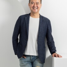 プロフィール写真 I様 2022.09.22 白いTシャツと紺色のジャケット、ダメージデニム姿の男性が、白い台に片手を置いて微笑むプロフィール撮影