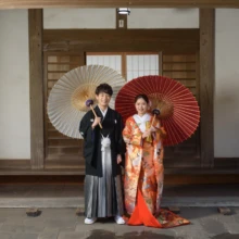 鎌倉 和装 K様・E様 2025.02.16 白と赤の番傘を背景に、赤い色打掛の新婦と黒紋付袴の新郎が並んで立つ鎌倉和装前撮り