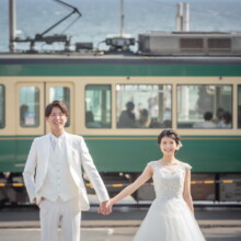 鎌倉 和洋装 K様・K様 2021.05.24 七里ヶ浜を走る緑色の江ノ電を背景に、手をつないで立つ白いウェディング衣装の新郎新婦の鎌倉フォトウェディング
