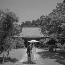 鎌倉 和洋装 K様・K様 2021.05.24 妙本寺の本堂と新緑が広がる境内で、白い和傘に入って並ぶ和装姿の新郎新婦をモノクロで捉えた一場面